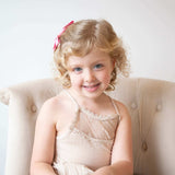 Adorable girl with curly hair wearing a pink grosgrain Kayla bow, smiling on a stylish chair, showcasing youthful charm.