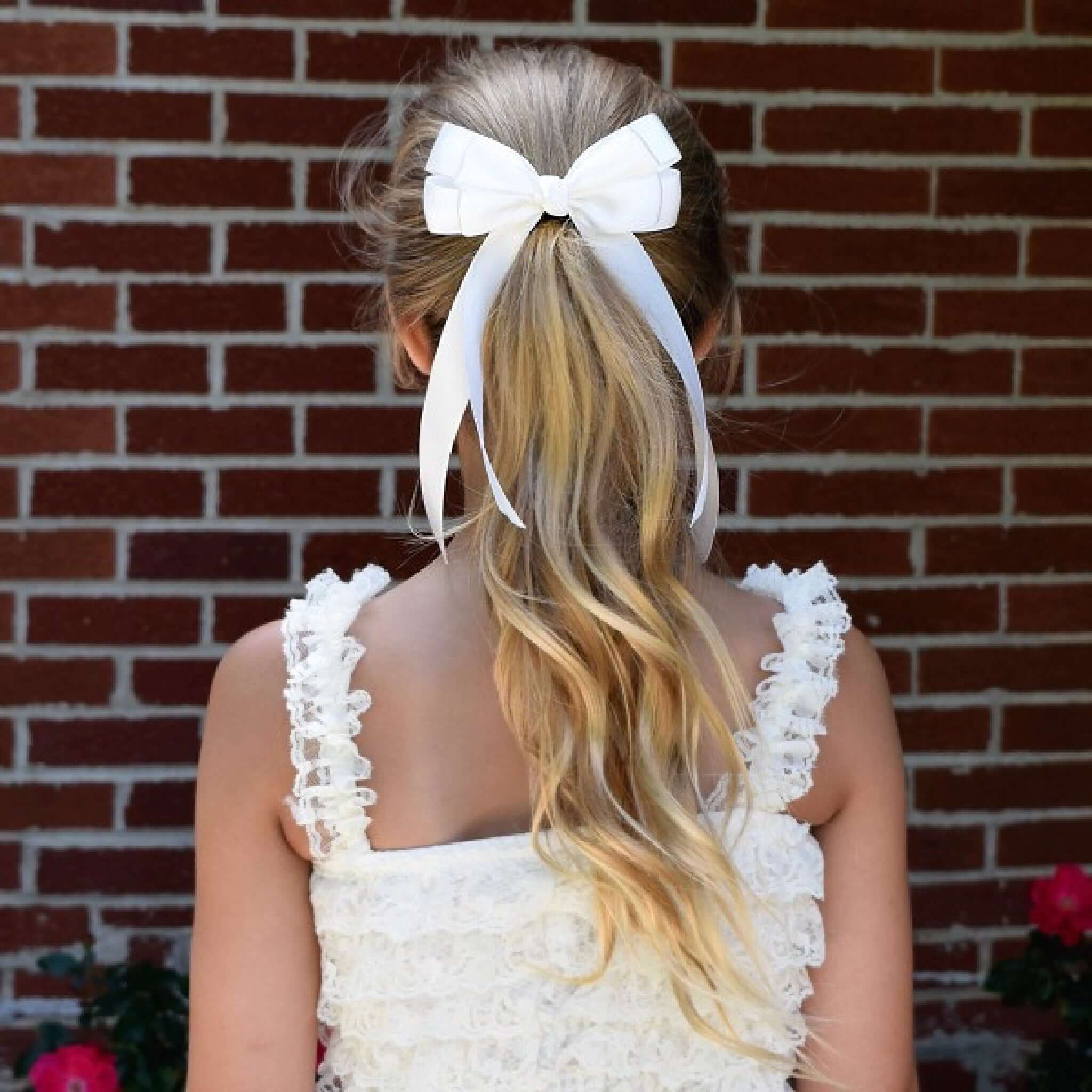 Girl with long blonde hair tied in a high ponytail with a ivory Emerson bow, wearing a white dress, against a brick wall.