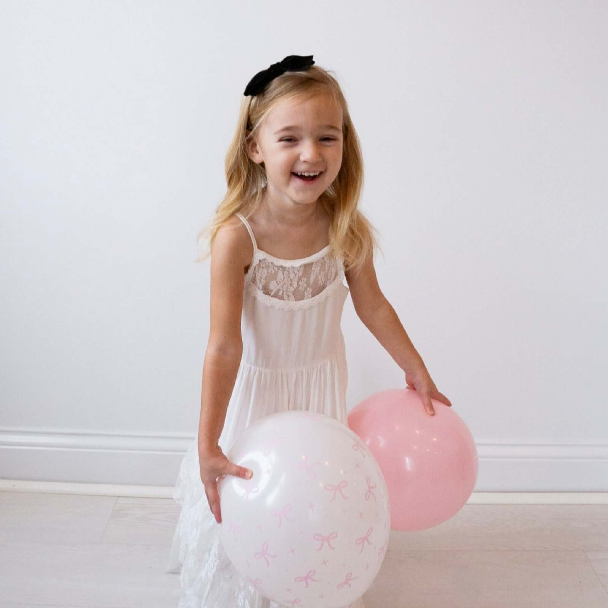 Young girl in a white dress holding balloons against a plain background