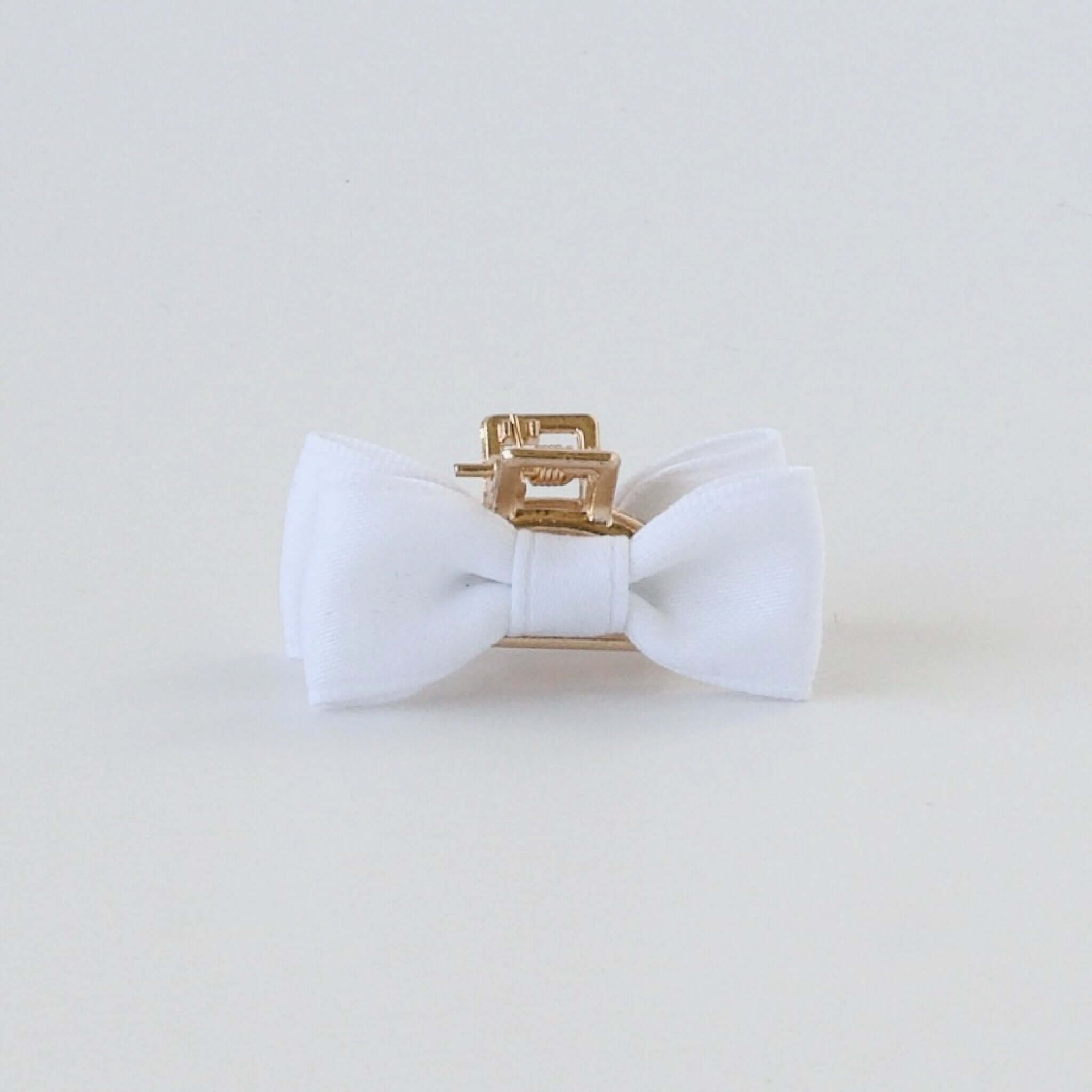 A white fabric bow tie with smooth texture, attached to a small gold hair clip, centered on a plain white background, exuding elegance and simplicity.