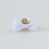 A white fabric bow tie with smooth texture, attached to a small gold hair clip, centered on a plain white background, exuding elegance and simplicity.