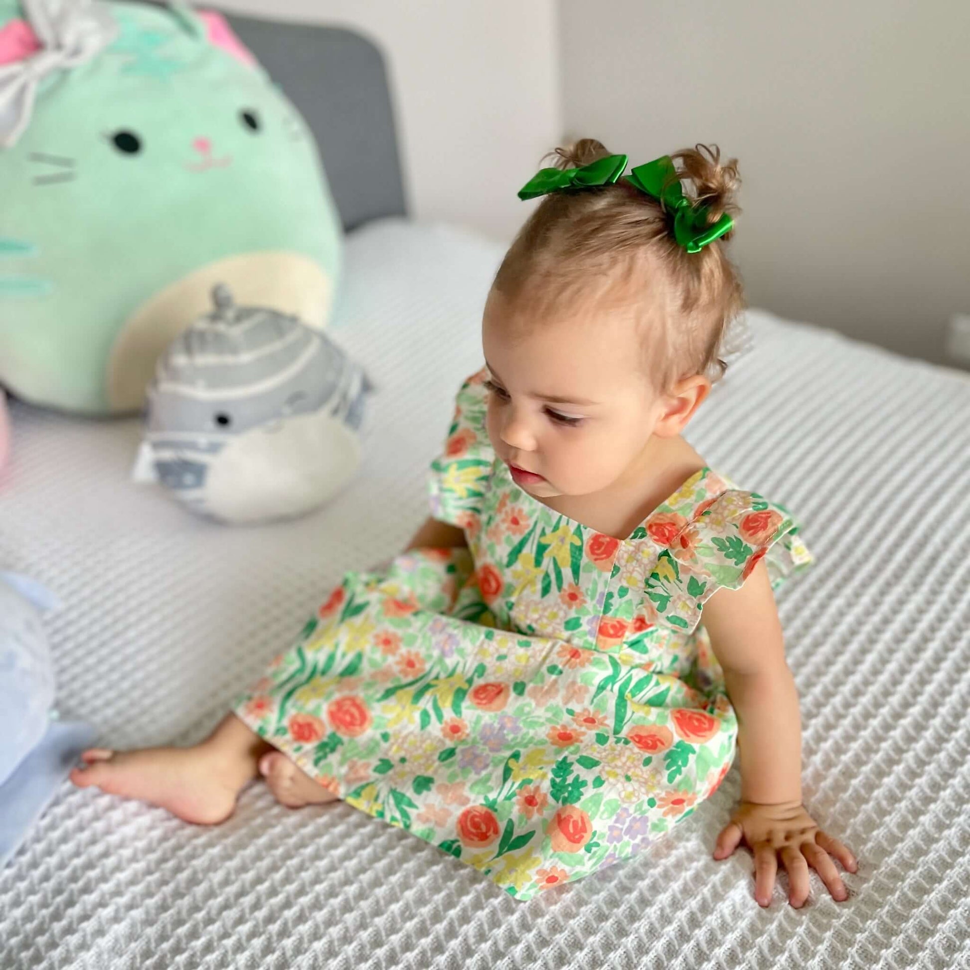 Baby wearing a floral dress and green hair bows sitting on a bed surrounded by plush toys
