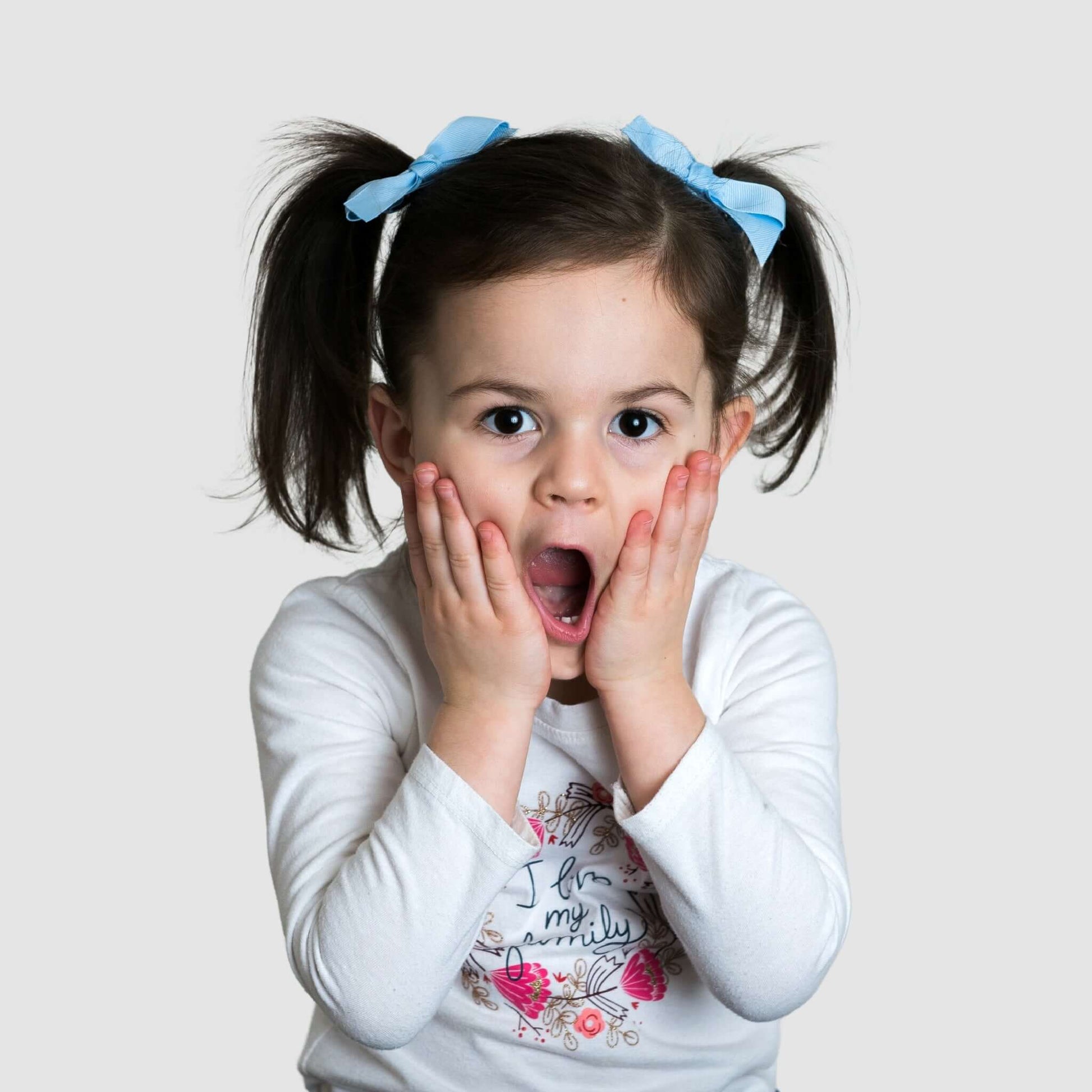 Toddler with pigtails wearing blue grosgrain baby Kayla bow clips, expressing surprise with hands on cheeks.