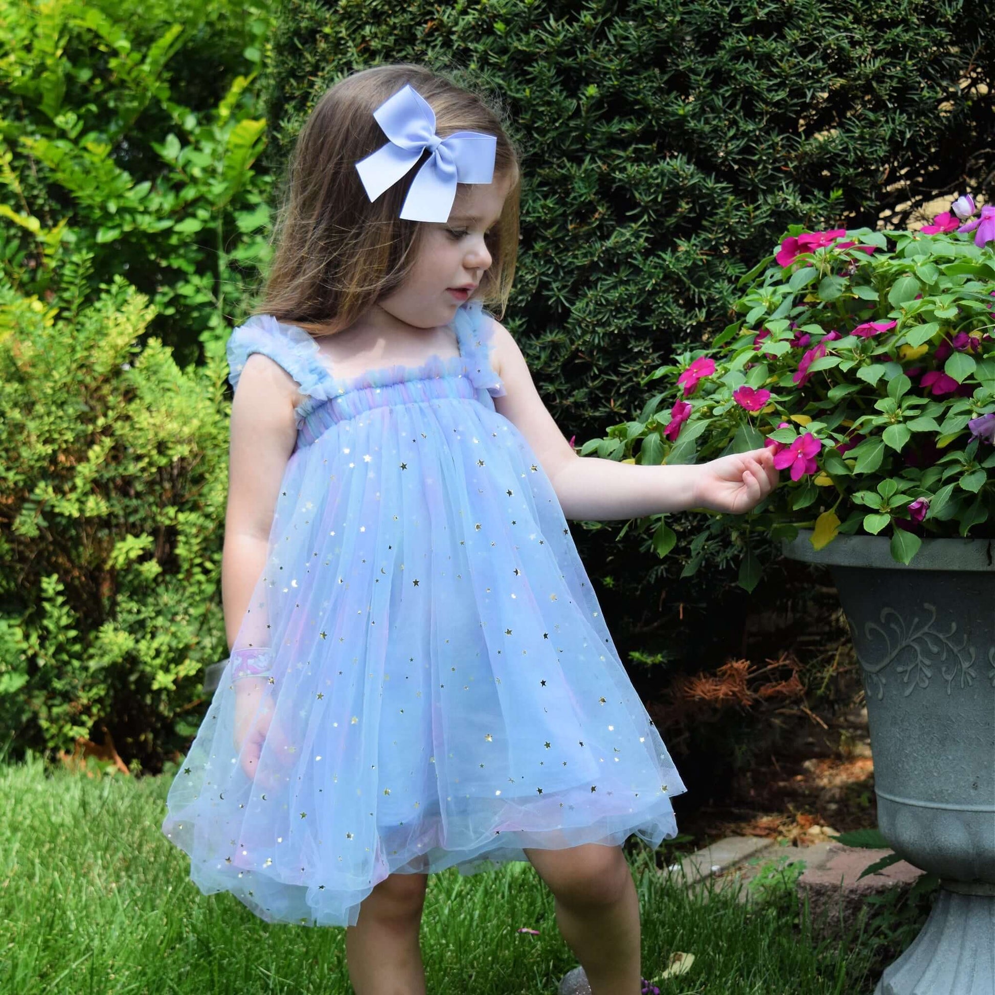 Toddler girl wearing a light blue dress with sparkles and a neutral 4 inch grosgrain sailor bow, standing next to a potted plant.