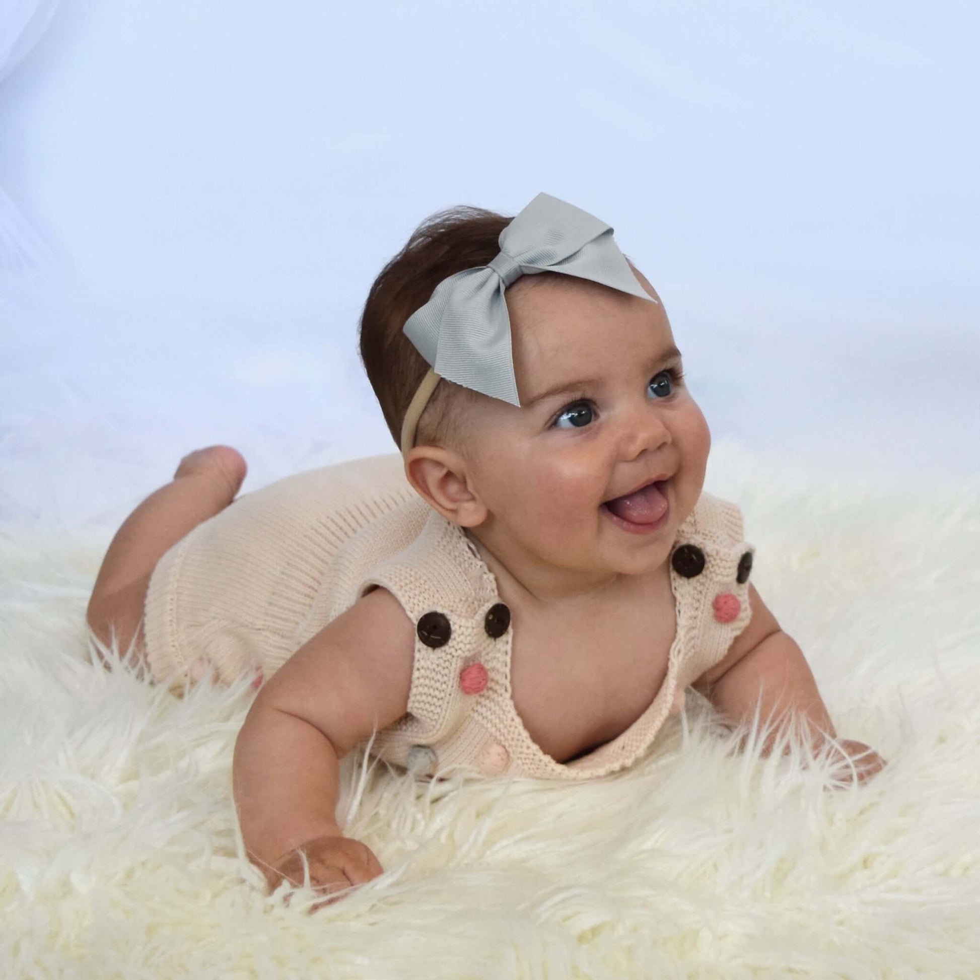 Baby wearing a gray 4 inch grosgrain bow headband, smiling while lying on fur rug, dressed in a cream knitted outfit with cute pom pom details.