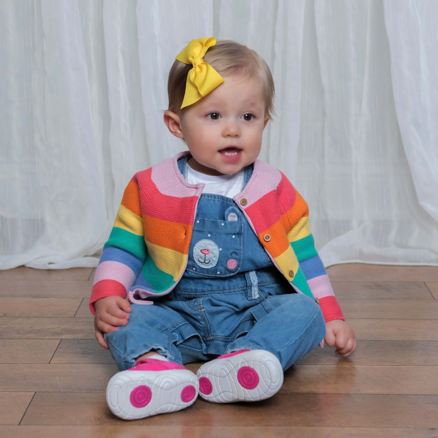 Toddler girl wearing a yellow bow clip, rainbow cardigan, and denim overalls, sitting on wooden floor