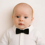 Baby wearing a white shirt and black velvet bow tie against a plain background