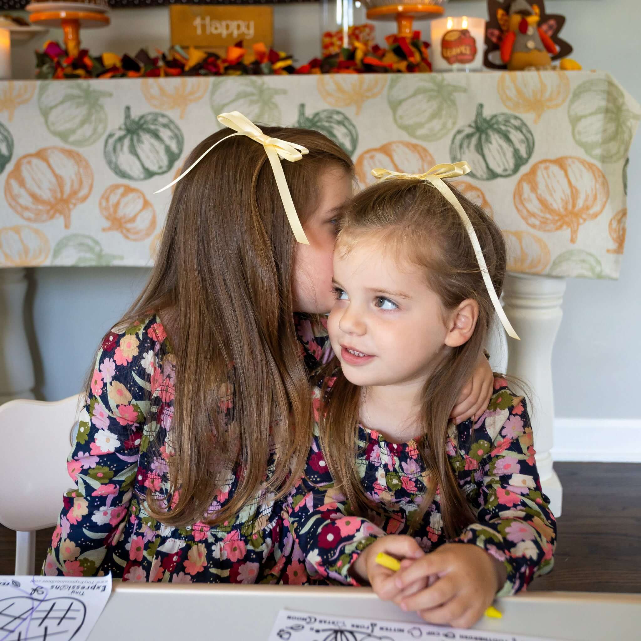 Two young girls in matching floral outfits with bows, sitting at a table with coloring books and crayons.