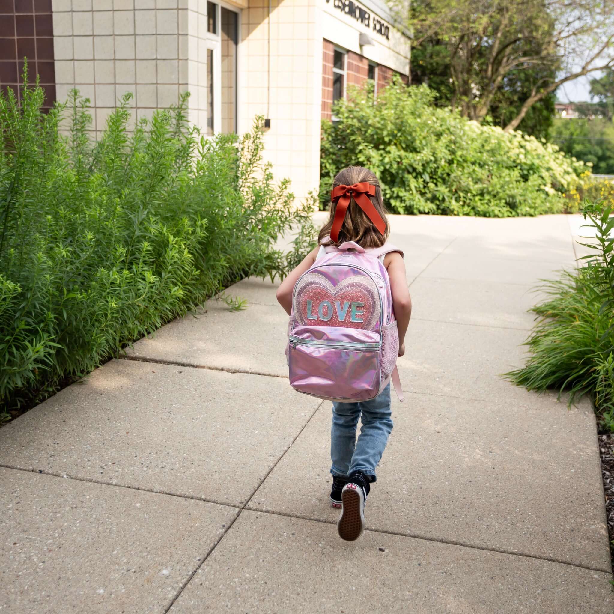 Child walking down a sidewalk with a pink backpack labeled 'LOVE' on a sunny day, wearing Grace grosgrain hair bow.