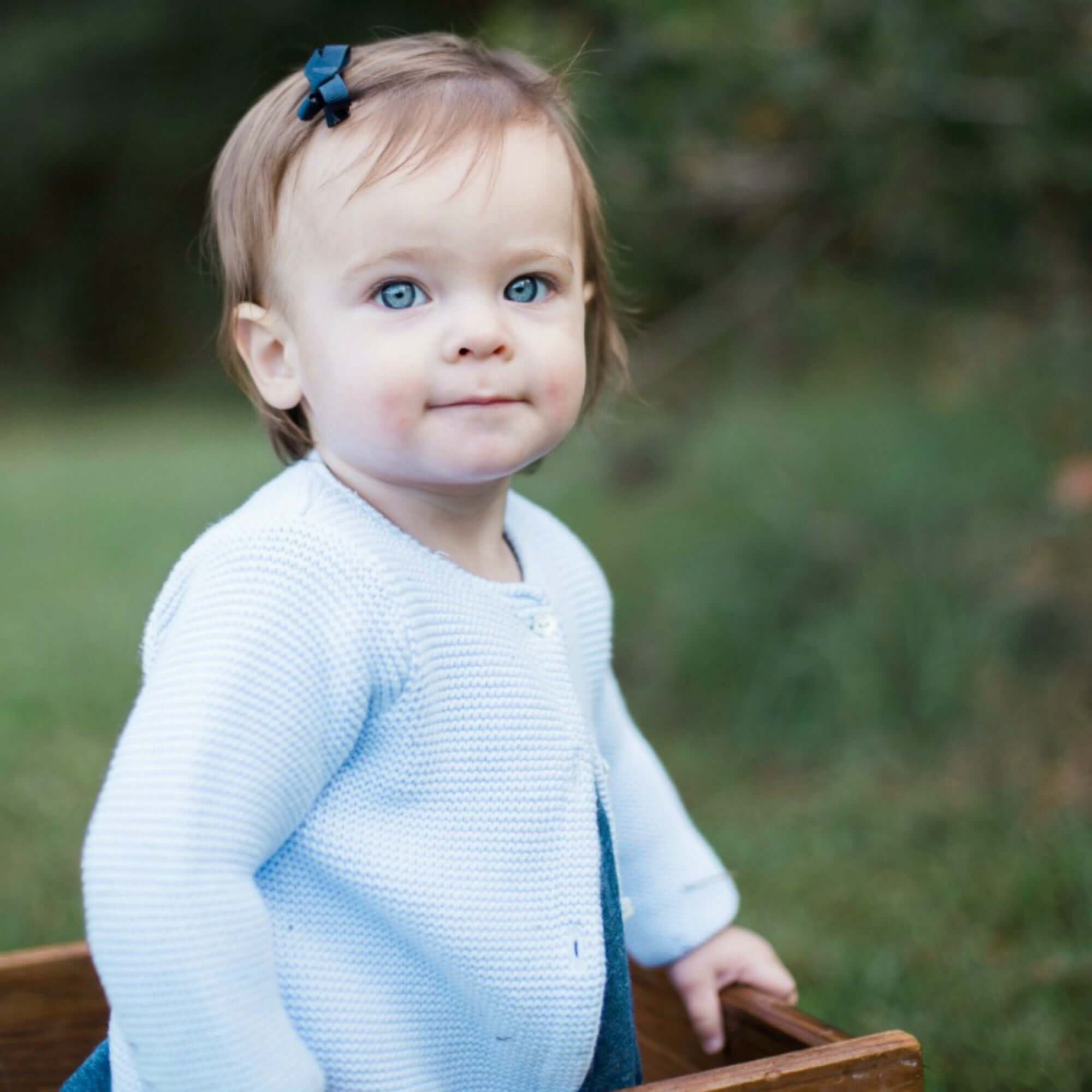 Baby in a light blue sweater standing outdoors with greenery in the background