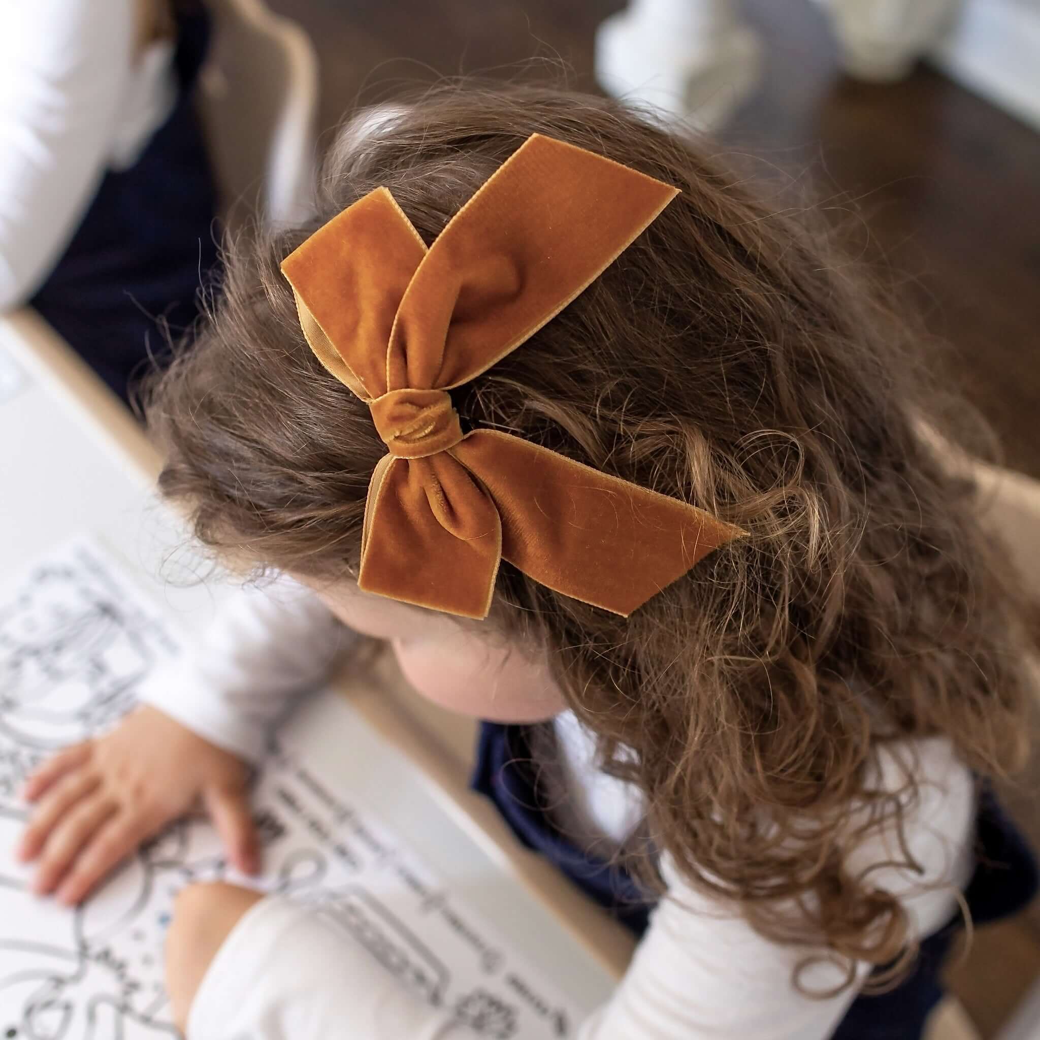 Child with brown hair wearing Flora velvet bow copper, sitting at a table.